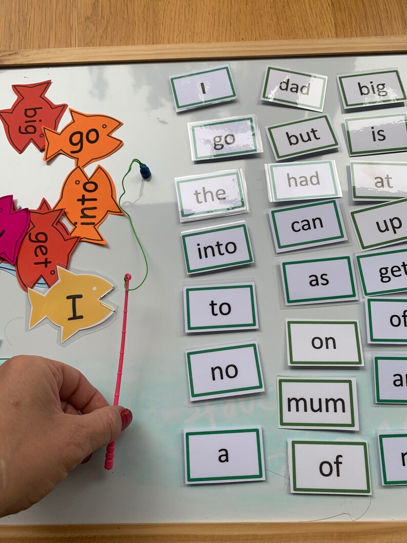 May include: A white board with a fishing rod and colourful fish with words on them. The words on the fish include "go", "into", "get", and "I". There are also word cards on the board with words like "dad", "big", "go", "but", "is", "had", "at", "can", "up", "get", "as", "to", "no", "a", "mum", "of", and "r".