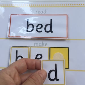 May include: A child's educational toy for learning to read. The word "bed" is printed on a white card with a red border. The letters "b", "e", and "d" are printed on separate cards with yellow borders. The letters are being placed in order to spell the word "bed".