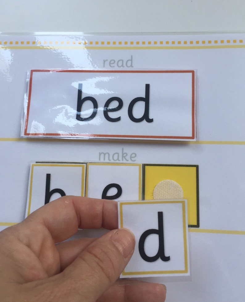 May include: A child's educational toy for learning to read. The word "bed" is printed on a white card with a red border. The letters "b", "e", and "d" are printed on separate cards with yellow borders. The letters are being placed in order to spell the word "bed".