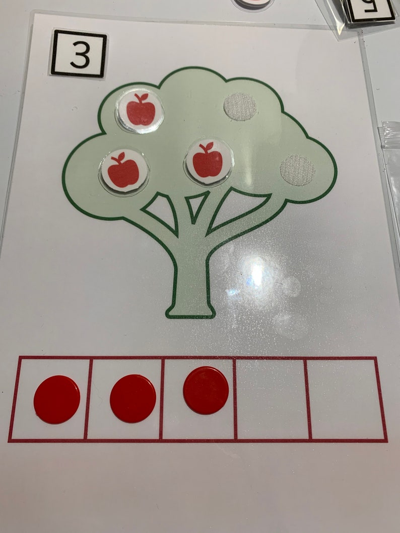 May include: A green tree with three red apples on it. The number 3 is written on a white square. There are three red circles in a row of five squares.