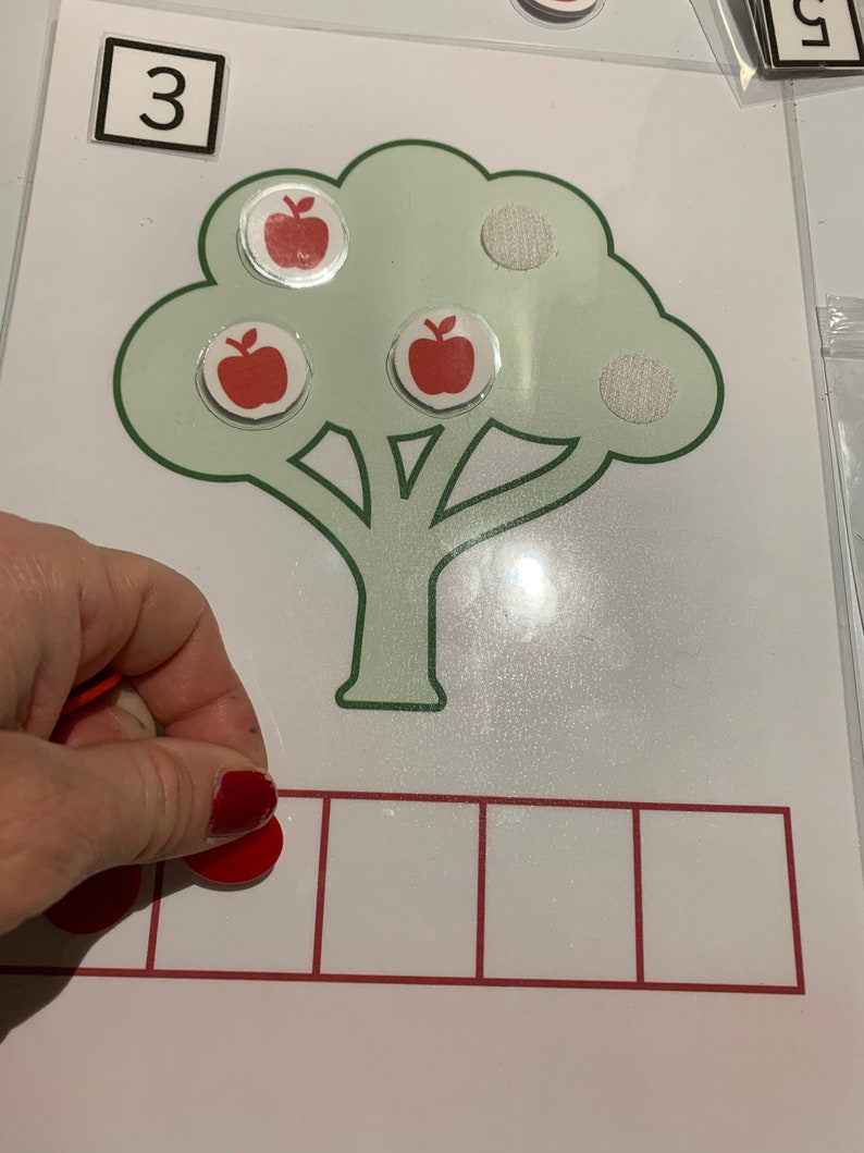 May include: A green tree with three red apples and three white circles. The number 3 is written in a black square at the top of the image. There are four empty red squares below the tree.