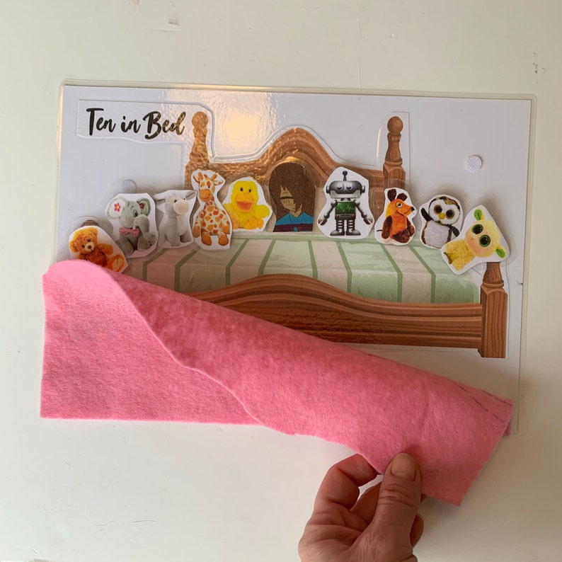 May include: A felt pink blanket is being pulled off of a wooden bed with a green and white patterned mattress. The bed has a headboard with a white background and the words "Ten in Bed" printed in black. There are ten felt animal characters on the bed, including a giraffe, a duck, a robot, and a dog.