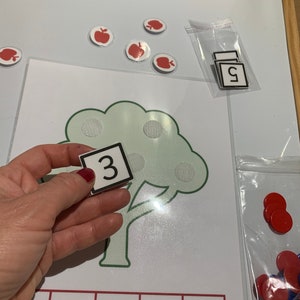 May include: A green tree with red apple stickers on the branches. A white card with the number "3" is held in front of the tree. There are red and blue counters in a plastic bag.