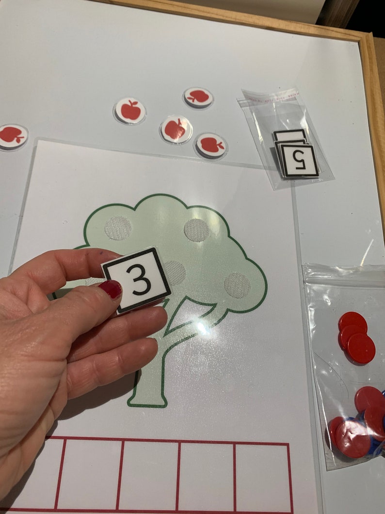 May include: A green tree with red apple stickers on the branches. A white card with the number "3" is held in front of the tree. There are red and blue counters in a plastic bag.