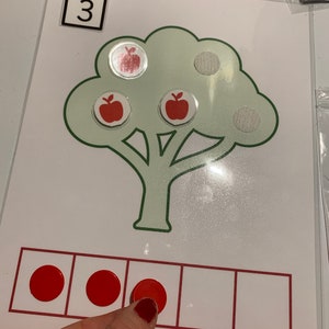 May include: A green tree with three red apples on it. The number 3 is written in a white square at the top of the image. There are three red circles in a row of five squares.