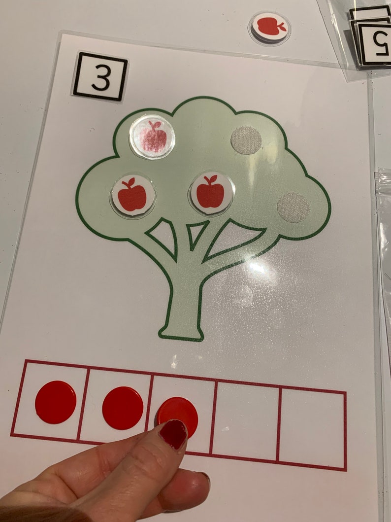 May include: A green tree with three red apples on it. The number 3 is written in a white square at the top of the image. There are three red circles in a row of five squares.