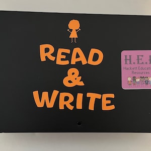May include: Black sign with orange text that reads "READ & WRITE" with a stick figure of a person with curly hair at the top and a pink square with the text "H.E.R. Hackett Educational Resources" and a group of stick figures at the bottom.