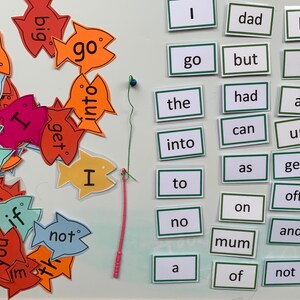 May include: A set of colourful fish-shaped cards with words printed on them, such as "go", "big", "not", "I", and "get".  The cards are arranged in a pile on a white surface.  There is a green and pink string attached to a blue bead, and a set of white cards with green borders, each with a single word printed on them, such as "dad", "the", "into", "to", "no", "mum", "of", "not", and "got".