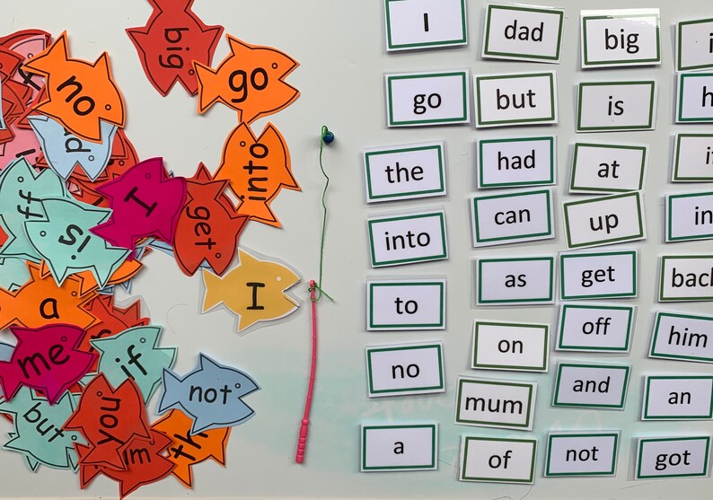 May include: A set of colourful fish-shaped cards with words printed on them, such as "go", "big", "not", "I", and "get".  The cards are arranged in a pile on a white surface.  There is a green and pink string attached to a blue bead, and a set of white cards with green borders, each with a single word printed on them, such as "dad", "the", "into", "to", "no", "mum", "of", "not", and "got".