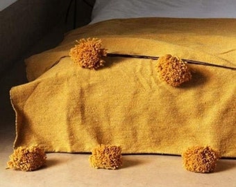 May include: A mustard yellow throw blanket with fluffy pom poms on a bed.