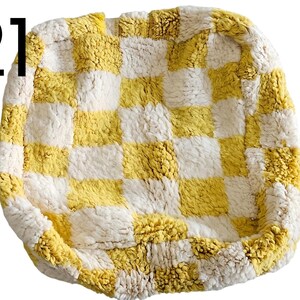 Puede incluir: Una cama cuadrada y afelpada para mascotas con un patr&oacute;n de tablero de ajedrez amarillo y blanco. La cama tiene un aspecto suave y texturizado y un borde elevado. El n&uacute;mero "21" es visible.