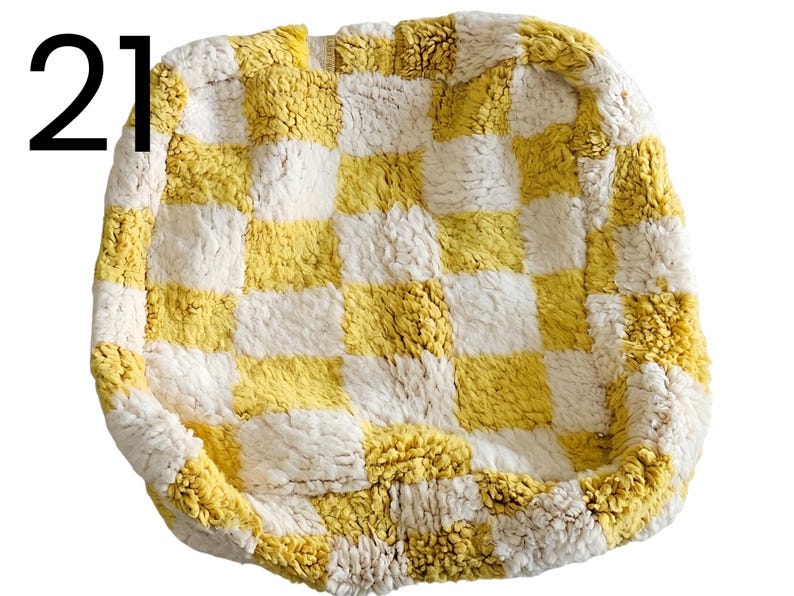 Puede incluir: Una cama cuadrada y afelpada para mascotas con un patr&oacute;n de tablero de ajedrez amarillo y blanco. La cama tiene un aspecto suave y texturizado y un borde elevado. El n&uacute;mero "21" es visible.