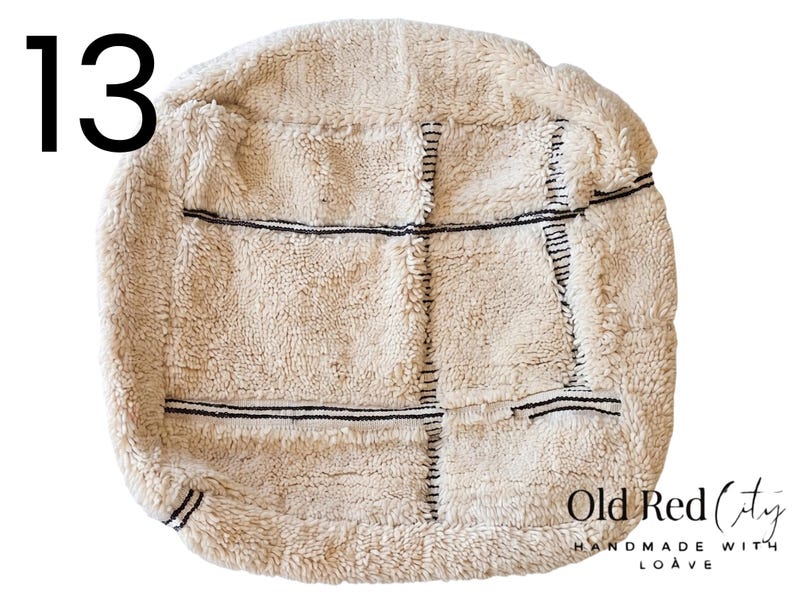 Puede incluir: Puf texturizado de color crema con l&iacute;neas cosidas negras. El puf tiene forma cuadrada con esquinas redondeadas. El n&uacute;mero 13 est&aacute; en la esquina superior izquierda. Las palabras "Old Red City" y "Handmade with Loave" est&aacute;n en la esquina inferior derecha.