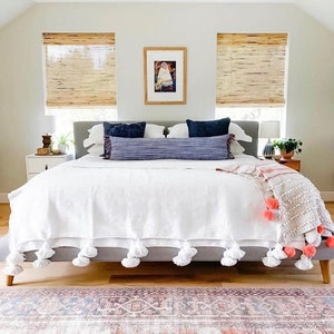 May include: A white bed with a white pom-pom trim blanket and a pink and white throw blanket. The bed has a gray headboard and two blue pillows. The bed is in a bedroom with a rug on the floor and two windows with bamboo blinds.