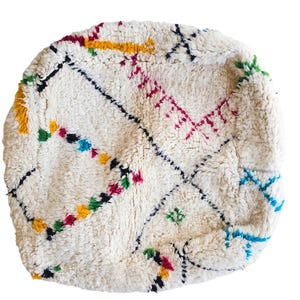 Puede incluir: Puf marroqu&iacute; redondo, blanquecino, con bordados geom&eacute;tricos coloridos. El dise&ntilde;o incluye detalles en negro, amarillo, rojo, verde y azul. El puf tiene un aspecto texturizado y tejido, ideal para sentarse o decorar.