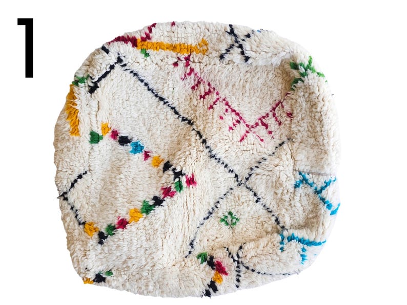Puede incluir: Puf marroqu&iacute; redondo, blanquecino, con bordados geom&eacute;tricos coloridos. El dise&ntilde;o incluye detalles en negro, amarillo, rojo, verde y azul. El puf tiene un aspecto texturizado y tejido, ideal para sentarse o decorar.