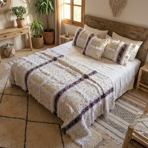 May include: A bed with a cream-colored blanket and pillows. The blanket has fringed edges and is accented with dark red and white patterned bands. The pillows have a similar design. The bed is set against a wooden headboard.