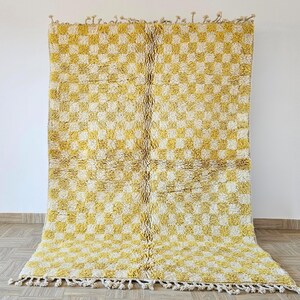 May include: A large, rectangular rug with a yellow and white chequered design. The rug has a shaggy texture and a fringe along the bottom edge. It is displayed against a white wall and wooden floor.