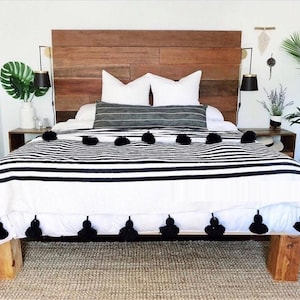 May include: A bed with a wooden headboard and a white and black striped blanket. The bed has white pillows and a gray lumbar pillow. Black tassels adorn the blanket's edge. The room is decorated with plants and a jute rug.