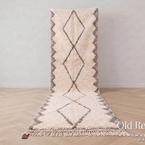Peut inclure: Un long tapis rectangulaire à poils longs de couleur crème, orné d'un motif répétitif de losanges gris foncé. Les bords présentent une bordure en zigzag et l'extrémité inférieure est agrémentée de franges en fils de couleur crème.