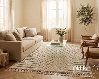 Op de afbeelding: Een woonkamer met neutrale tinten met een beige bank, een patroonkleed en twee fauteuils. Een glazen salontafel en een decoratieve boom zijn ook aanwezig. De kamer heeft een neutraal kleurenpalet.