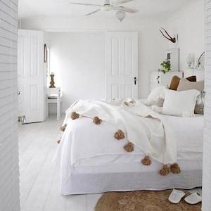 May include: A white bedroom with a white bed, white bedding, and a white throw blanket with brown tassels. The bed is against a white wall and there is a white door to the left of the bed. The floor is white wood.