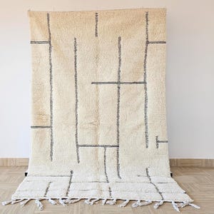 May include: Cream-colored, rectangular rug with a geometric design. The rug features vertical and horizontal dark gray lines creating a grid-like pattern. The bottom edge has a fringe detail, adding texture and visual interest. This rug is suitable for living rooms and bedrooms.
