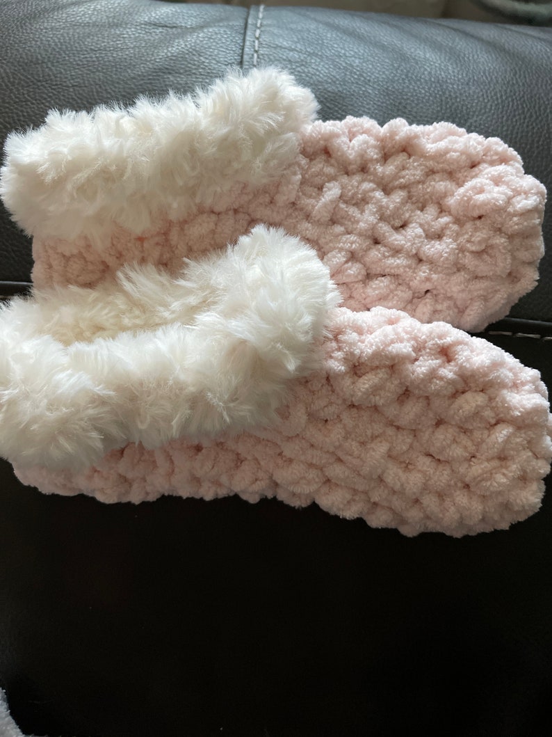 May include: A pair of pink fuzzy slippers with white faux fur trim. The slippers are crocheted and have a soft, cozy texture.