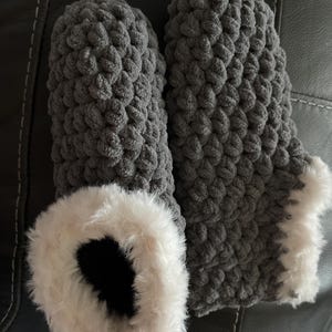 May include: A pair of gray knitted slippers with a white faux fur trim. The slippers are made with a chunky knit stitch.
