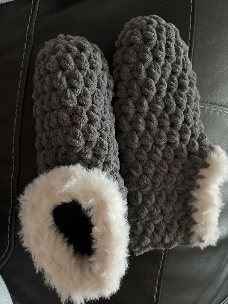 May include: A pair of gray knitted slippers with a white faux fur trim. The slippers are made with a chunky knit stitch.