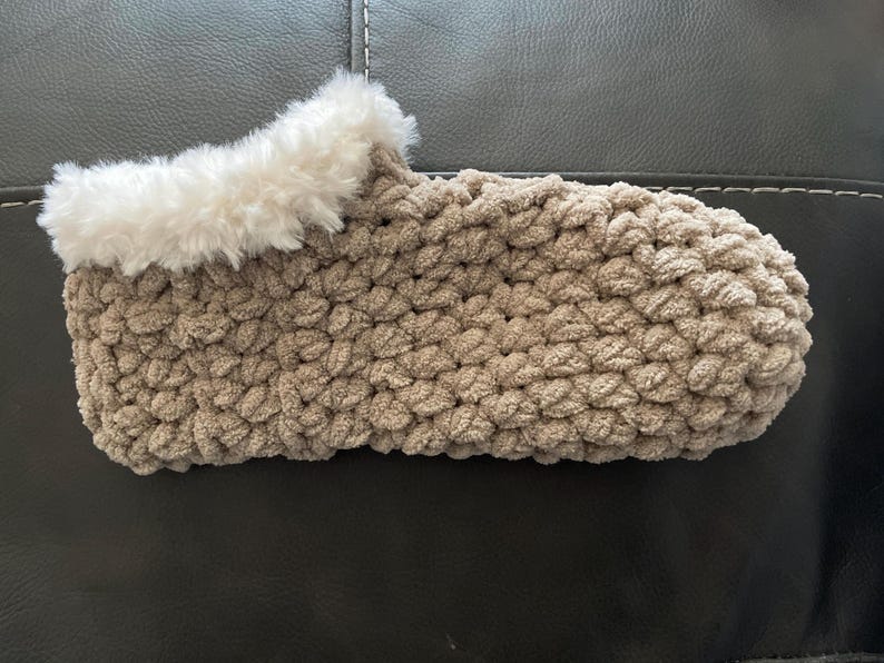 May include: A pair of cozy brown crocheted slippers with a white faux fur trim. The slippers are made with a chunky yarn and have a soft, comfortable texture.