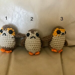 May include: Three crocheted stuffed toys resembling the Porgs from Star Wars. The toys are brown with orange feet and yellow accents around their eyes. The toys are numbered 1, 2, and 3.