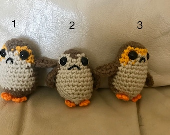 May include: Three crocheted stuffed toys resembling the Porgs from Star Wars. The toys are brown with orange feet and yellow accents around their eyes. The toys are numbered 1, 2, and 3.