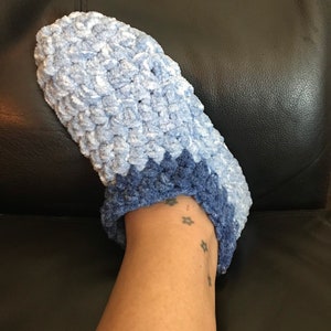 May include: A pair of blue crocheted slippers with a lighter blue top and a darker blue bottom. The slippers are worn on a person's foot.