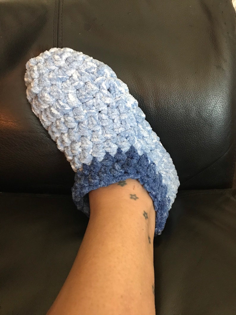 May include: A pair of blue crocheted slippers with a lighter blue top and a darker blue bottom. The slippers are worn on a person's foot.