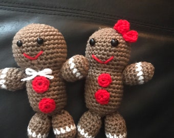 May include: Two crocheted gingerbread people, one with a red bow, standing on a black surface. They are brown with white trim and red buttons.
