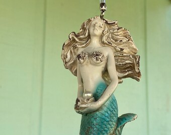 May include: A hanging mermaid wind chime with a starfish and crystal bead accents. The mermaid has long, flowing hair, seashell-style bra, and a teal tail. The starfish is white and textured. The wind chime is suspended by a clear string.