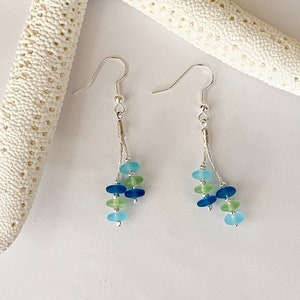 May include: A pair of dangle earrings featuring silver-toned hooks and thin chains. Each earring displays a cluster of small, smooth, rounded beads in shades of light blue, dark blue, and pale green, arranged in a vertical, multi-strand design.