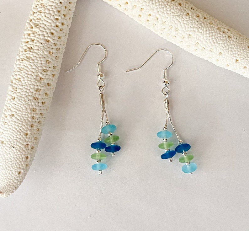 May include: A pair of dangle earrings featuring silver-toned hooks and thin chains. Each earring displays a cluster of small, smooth, rounded beads in shades of light blue, dark blue, and pale green, arranged in a vertical, multi-strand design.