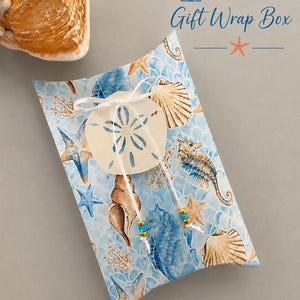 May include: A rectangular pillow box with a light blue background featuring a pattern of seashells, starfish, and seahorses. A white string ties a sand dollar cutout to the center, accented with small blue and gold beads. Text reads FREE Gift Wrap Box.