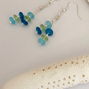 May include: A pair of dangle earrings featuring silver-toned hooks and chains. Each earring is adorned with multiple small, circular, frosted glass beads in shades of light blue, dark blue, and pale green, arranged in a cascading cluster design.