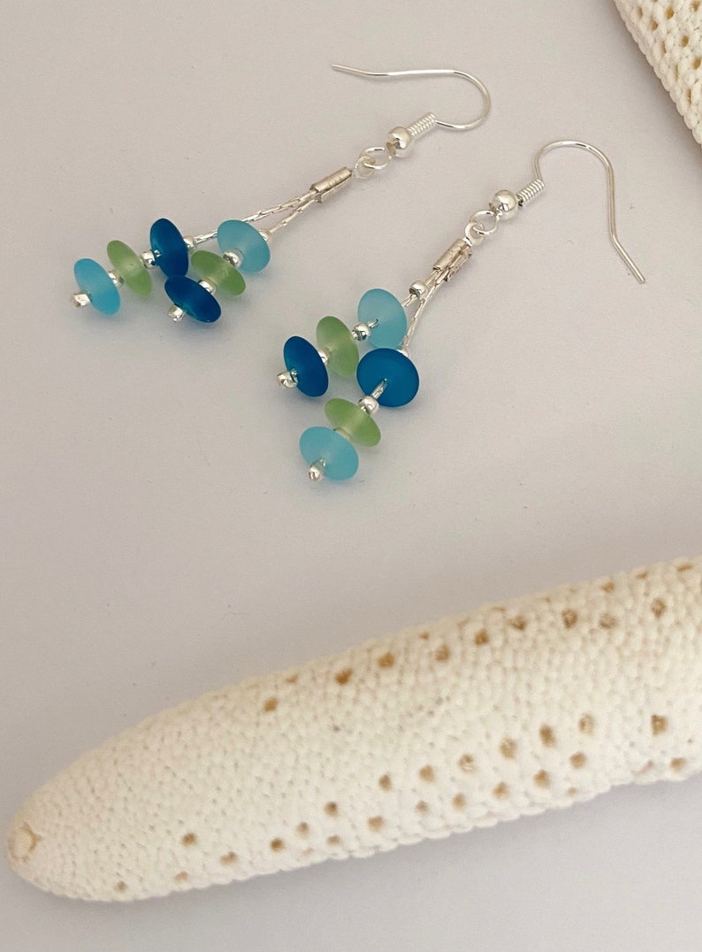 May include: A pair of dangle earrings featuring silver-toned hooks and chains. Each earring is adorned with multiple small, circular, frosted glass beads in shades of light blue, dark blue, and pale green, arranged in a cascading cluster design.