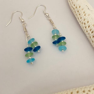May include: A pair of dangle earrings featuring silver-toned hooks and chains. Each earring is adorned with multiple small, smooth, frosted glass beads in shades of light blue, dark blue, and pale green, arranged in a cascading vertical design.