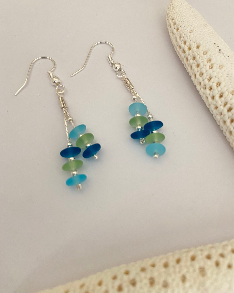 May include: A pair of dangle earrings featuring silver-toned hooks and chains. Each earring is adorned with multiple small, smooth, frosted glass beads in shades of light blue, dark blue, and pale green, arranged in a cascading vertical design.
