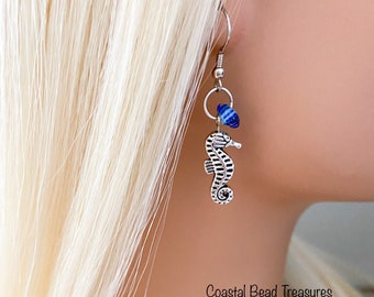 May include: Silver seahorse earrings with a blue and white bead accent. The earrings have a fish hook closure and a small ring connecting the seahorse charm and the bead. The text "Coastal Bead Treasures" is visible.