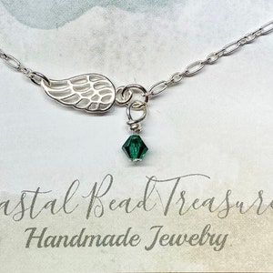 May include: A silver necklace featuring a delicate wing charm and a small, faceted, dark green crystal pendant. The necklace is displayed on a card with the text "Coastal Bead Treasures Handmade Jewelry".
