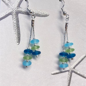 May include: A pair of dangle earrings featuring silver-toned hooks and double chains. Each earring displays small, frosted, sea-glass-style beads in shades of light blue, dark blue, and pale green, arranged in a tiered, multi-strand design.