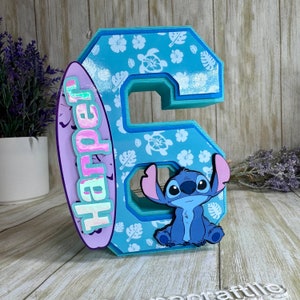 May include: A light blue number 6, decorated with a tropical pattern of flowers and turtles. A purple surfboard with the name "Harper" is attached to the side. A cartoon image of Stitch is on the front.