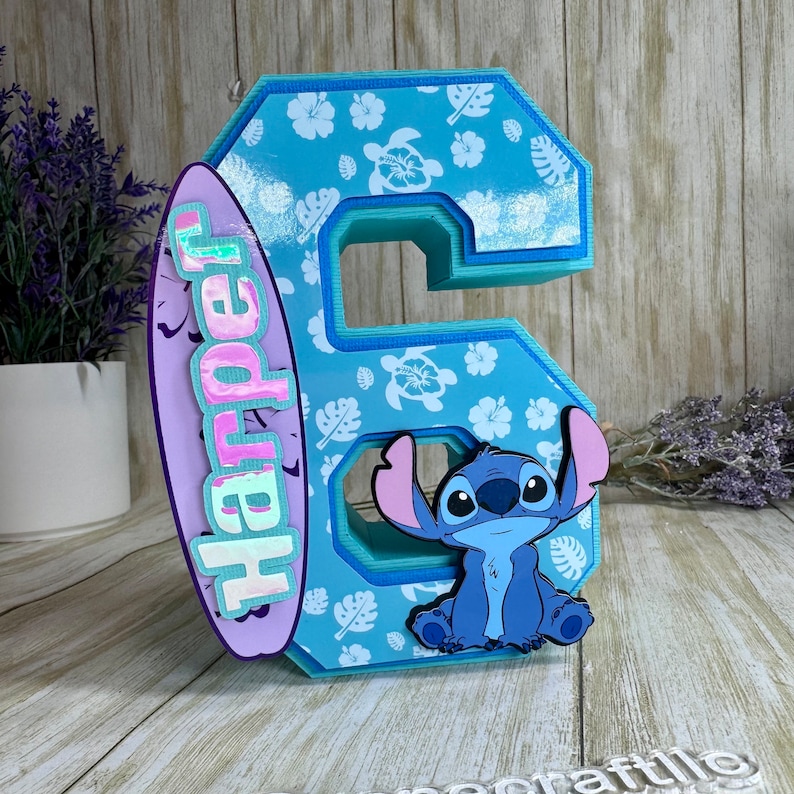 May include: A light blue number 6, decorated with a tropical pattern of flowers and turtles. A purple surfboard with the name "Harper" is attached to the side. A cartoon image of Stitch is on the front.
