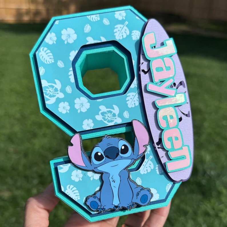 May include: A light blue number nine decoration with a Stitch character and a surfboard with the name "Jayleen". The number is decorated with white floral patterns. The surfboard is purple with pink and green accents.